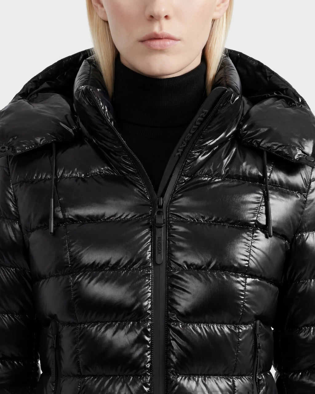 A person with light blonde hair wears the Rudsak MELISSA C - 8125695 Black women's hooded puffer jacket, partially zipped over a black turtleneck. The quilted, lightweight down design reveals only the lower half of her face.