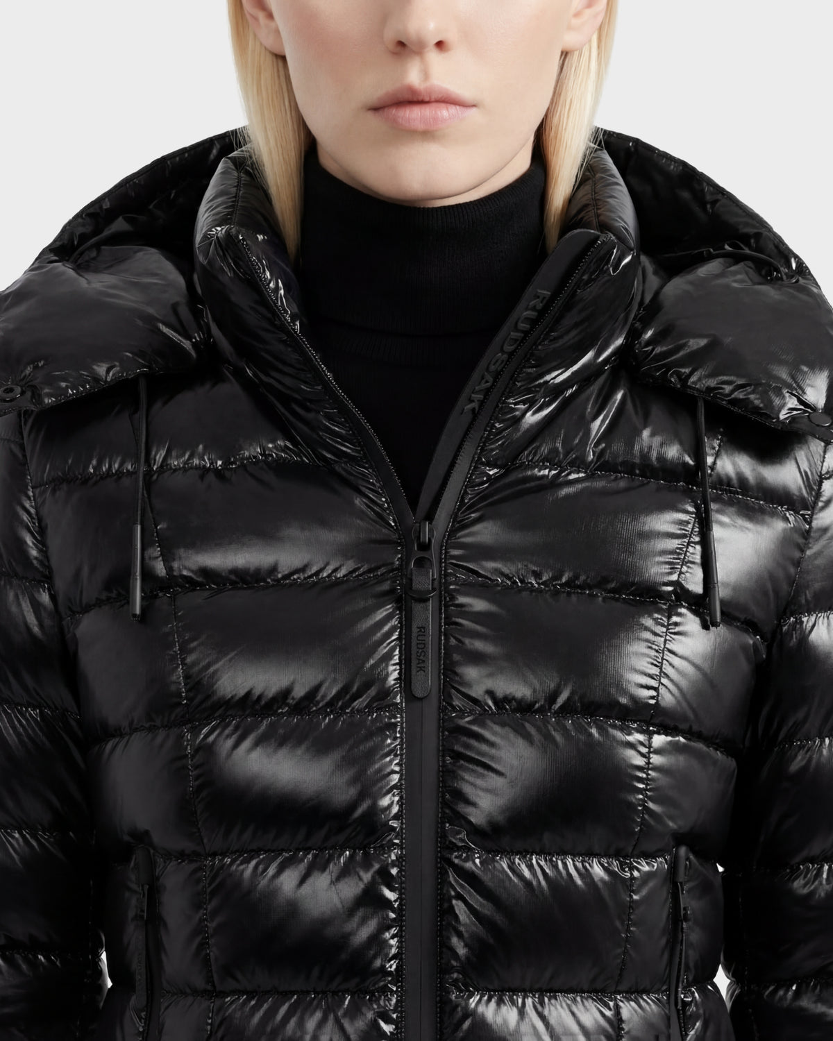 A person with light blonde hair wears the Rudsak MELISSA C - 8125695 Black women's hooded puffer jacket, partially zipped over a black turtleneck. The quilted, lightweight down design reveals only the lower half of her face.