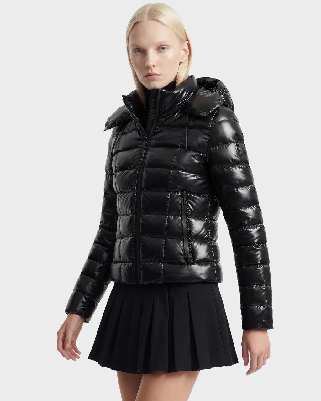 A woman with straight blonde hair wears the Rudsak MELISSA C - 8125695 Black puffer jacket with a high collar and a short black pleated skirt, posing against a plain light gray background.