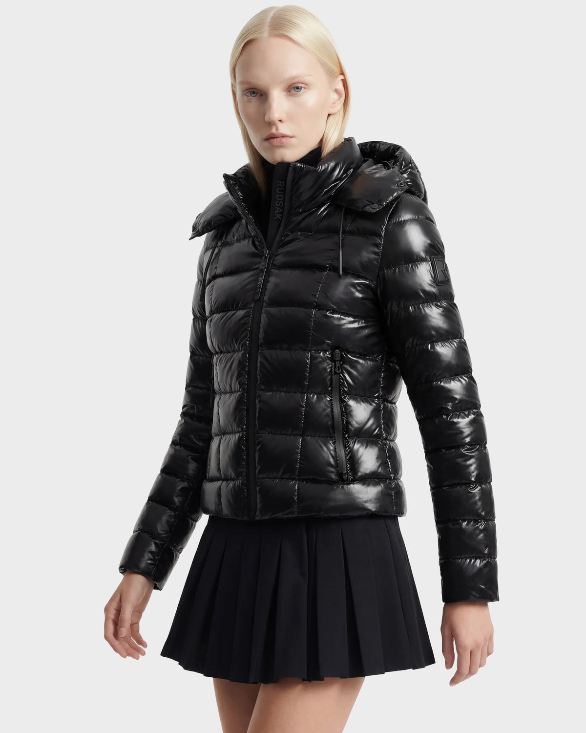 A woman with straight blonde hair wears the Rudsak MELISSA C - 8125695 Black puffer jacket with a high collar and a short black pleated skirt, posing against a plain light gray background.