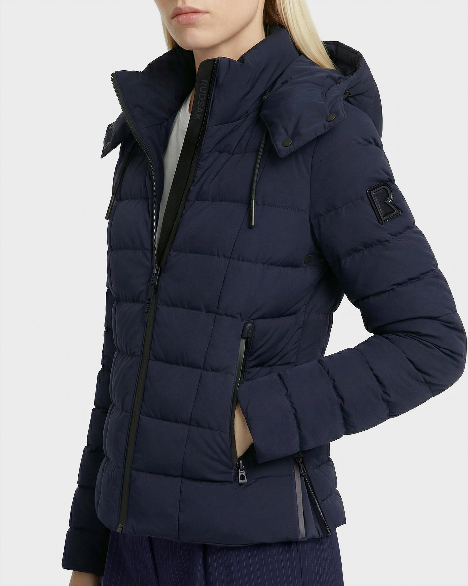 A woman stands with one hand in her pocket, wearing the Rudsak MELISSA - 8125054 Navy women’s puffer jacket with a hood, zippered pockets, and sleeve patch. She pairs it with dark pants; her face is partially out of frame.