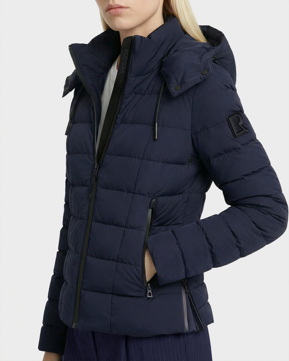 A woman stands with one hand in her pocket, wearing the Rudsak MELISSA - 8125054 Navy women’s puffer jacket with a hood, zippered pockets, and sleeve patch. She pairs it with dark pants; her face is partially out of frame.