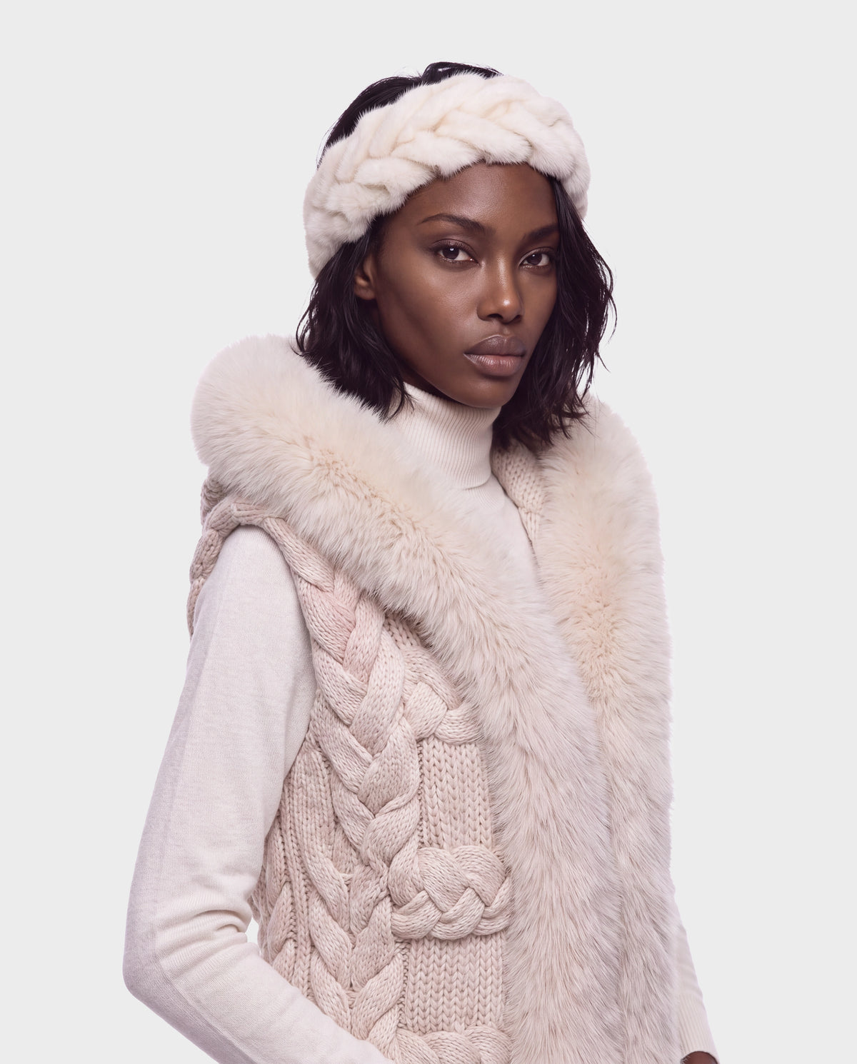 A woman wearing a cream-colored Rudsak MILA - 8426518 Vanilla knit vest with a fur-trimmed hood and a matching braided headband over a turtleneck sweater.