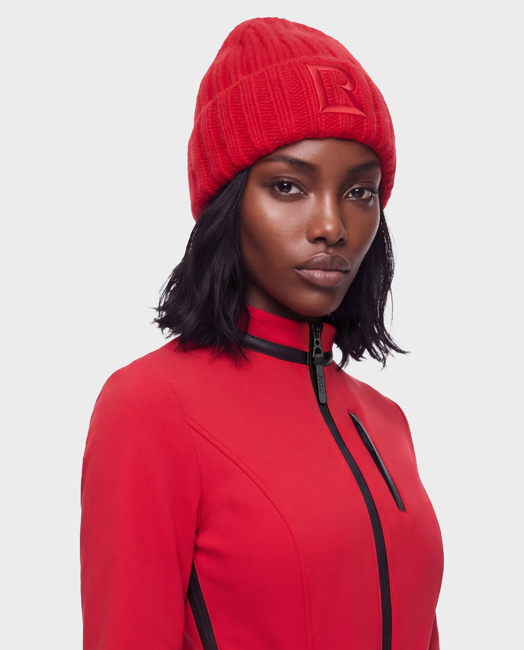 A person wearing a bright red ribbed knit MISSA - 8426512 Flame Red beanie with an embossed letter R on the front and a matching red zip-up jacket.