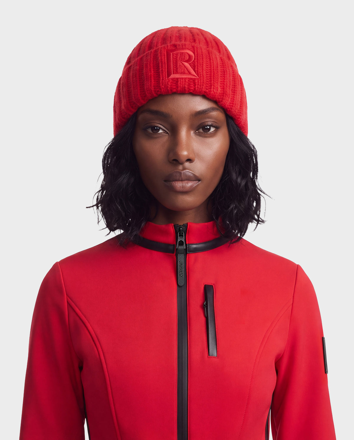 A person wearing a bright red Rudsak MISSA - 8426512 Flame Red ribbed knit beanie with an R on the front and a matching red zip-up jacket with black trim.