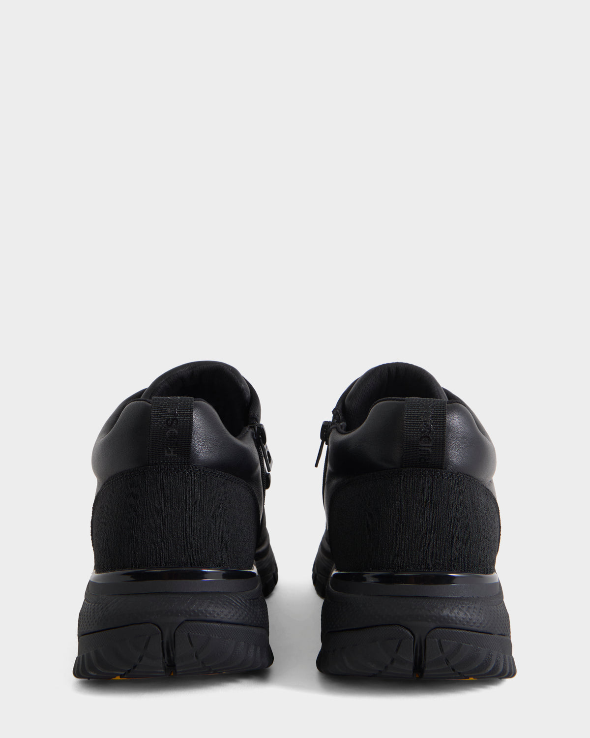 A pair of Rudsak MAX - 6226502 Black men’s sneakers is shown from the back against a plain light gray background. These utility-inspired athletic shoes feature textured soles and uppers made from a combination of smooth and knit fabrics.