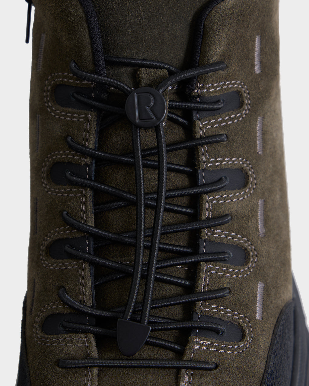 Close-up of the Rudsak MAX S - 6226505 Olive, a brown suede boot with black elastic laces threaded through black eyelets, spotlighting a round button with the letter R at the center and visible white stitching along the sides—this trail-inspired design seamlessly fuses rugged durability with contemporary men’s sneaker style.