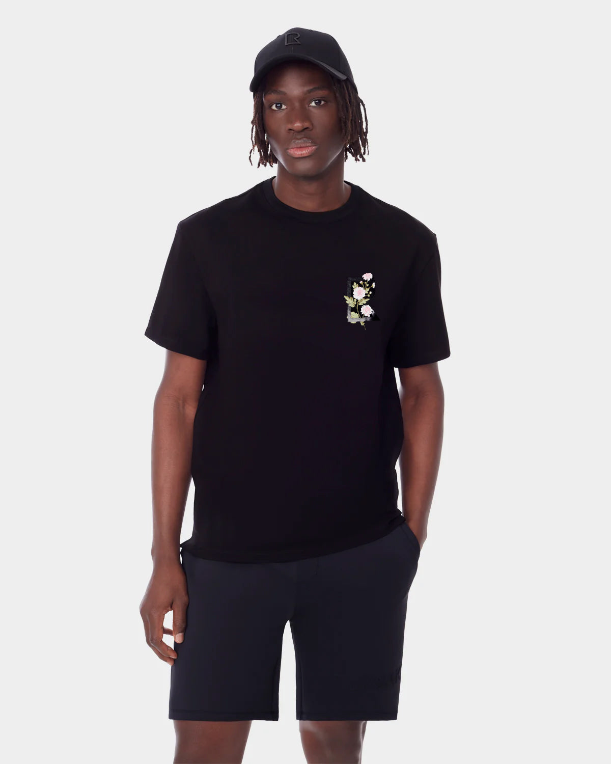 Wearing a black cap, unisex fit DIAR - 9523003 M BLACK T-shirt by Rudsak with a small floral chest design and black shorts, someone stands against a plain light background—an elevated everyday look.