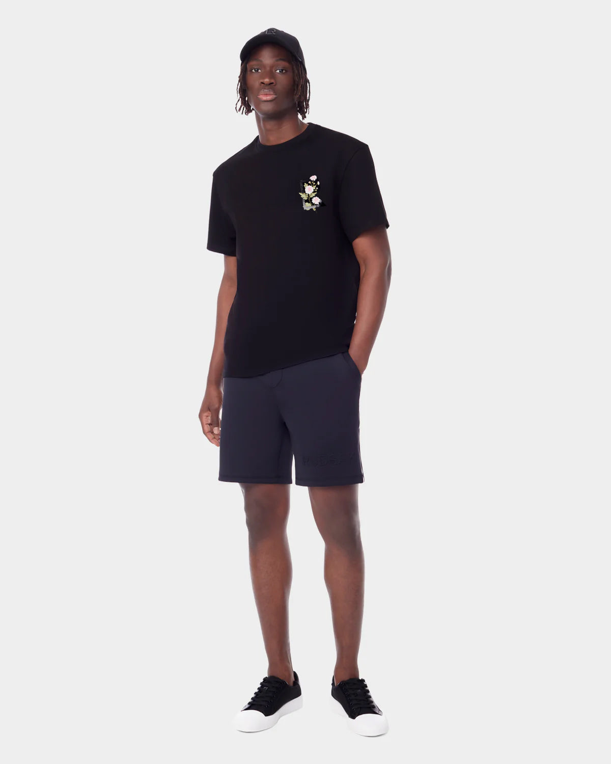 A man models a sleek everyday look in unisex style: black cap, Rudsak DIAR - 9523003 M BLACK T-shirt with small floral detail, black shorts, and black sneakers with white soles against a plain white backdrop.