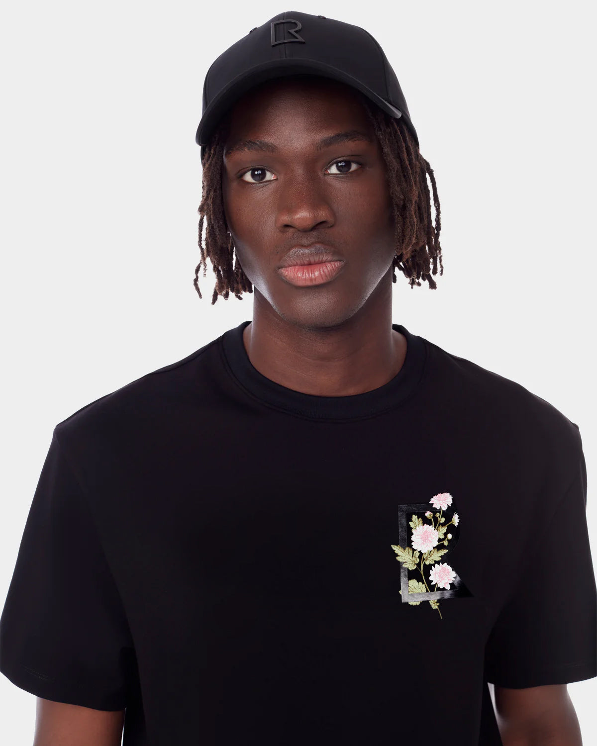 A person wearing the Rudsak DIAR - 9523003 M BLACK T-shirt with a floral graphic and a black baseball cap stands against a white background, showcasing an elevated everyday style while looking at the camera.