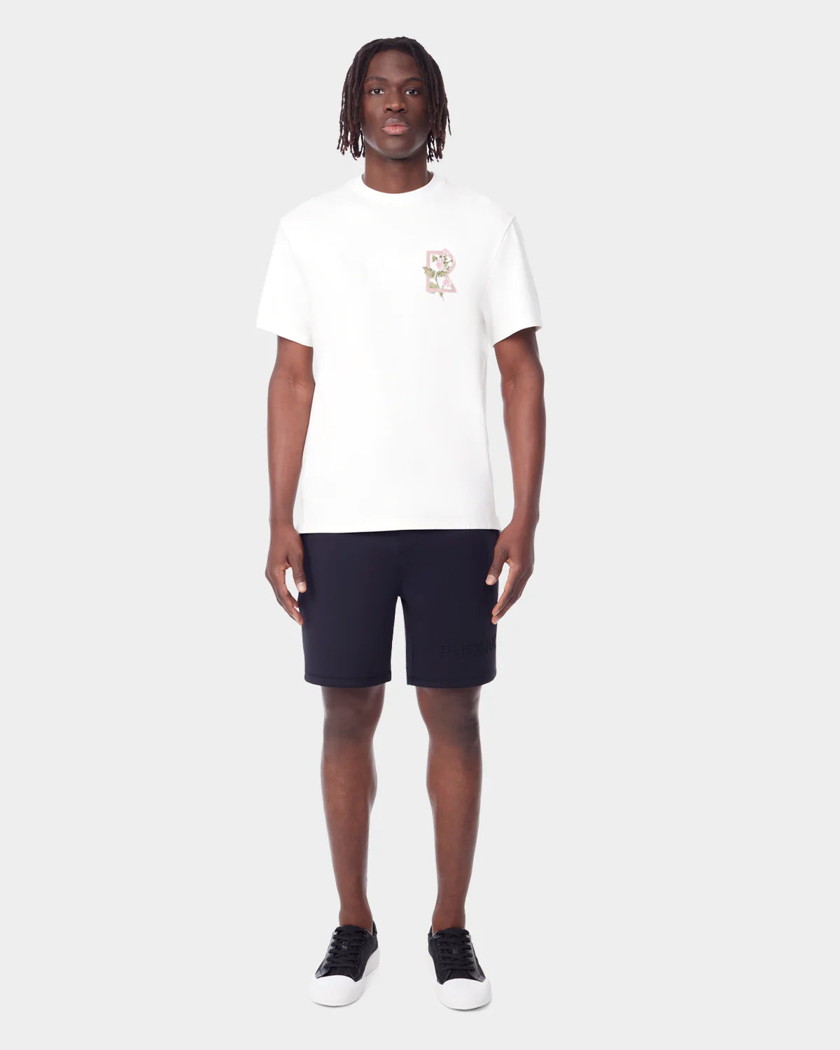 A man with short dreadlocks wears the Rudsak DIAR - 9523003 M OPTIC WHITE unisex T-shirt with a small chest graphic, paired with black shorts and black-and-white sneakers, standing against a plain light gray background.