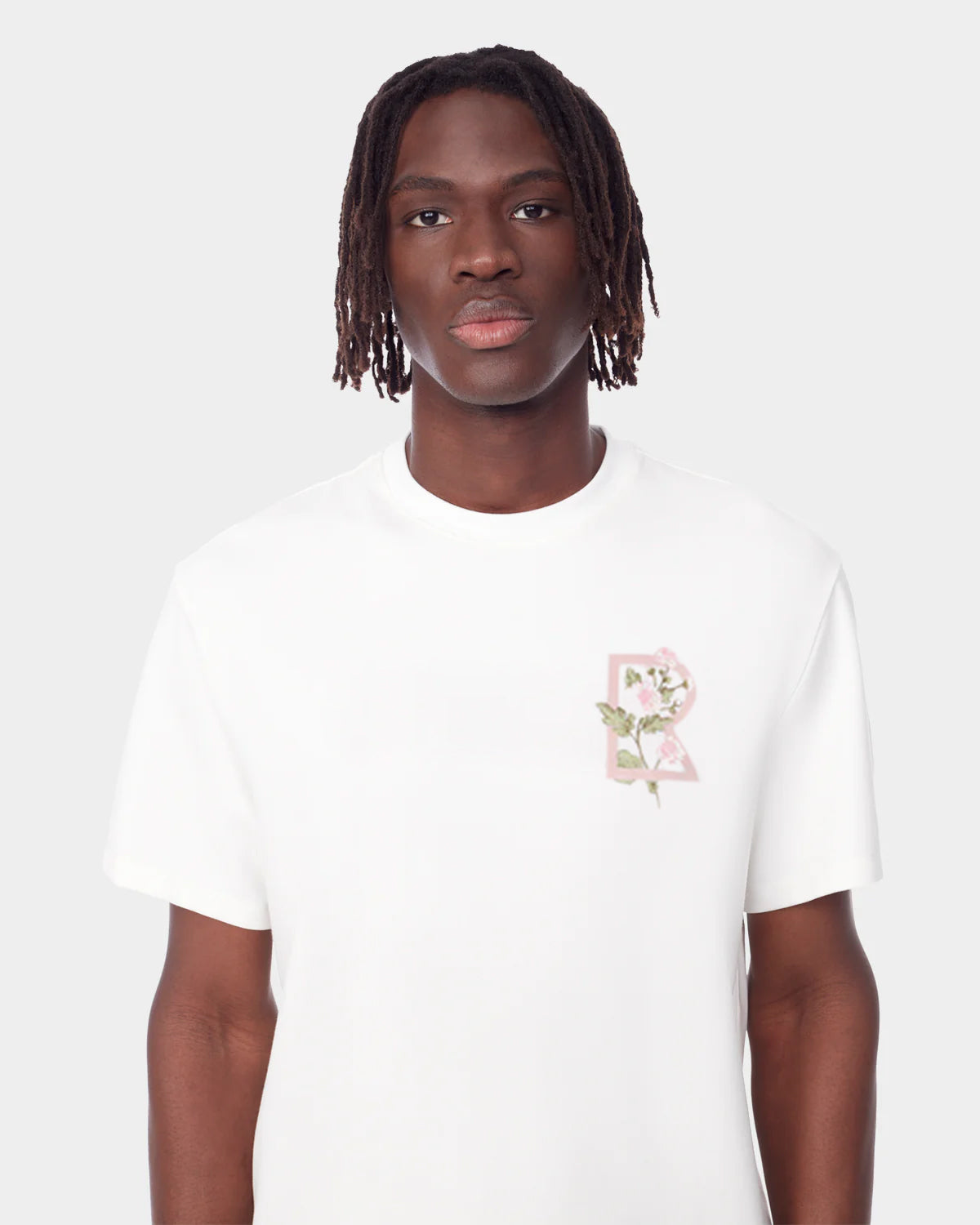 A person with medium-length dreadlocks wears the Rudsak DIAR - 9523003 M OPTIC WHITE t-shirt, featuring a floral letter R design and classic cut, standing against a light gray background.