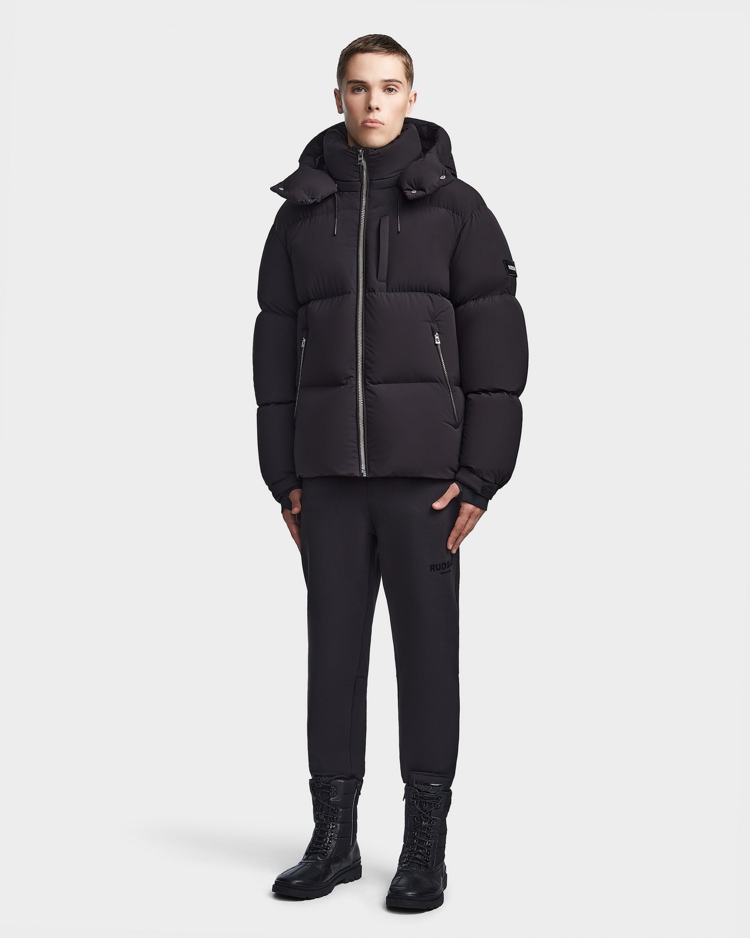 A person stands against a white background wearing the Rudsak VINCENT M - 6123510 Black men’s down puffer jacket, paired with black pants and black lace-up boots, looking straight ahead with hands by their sides.