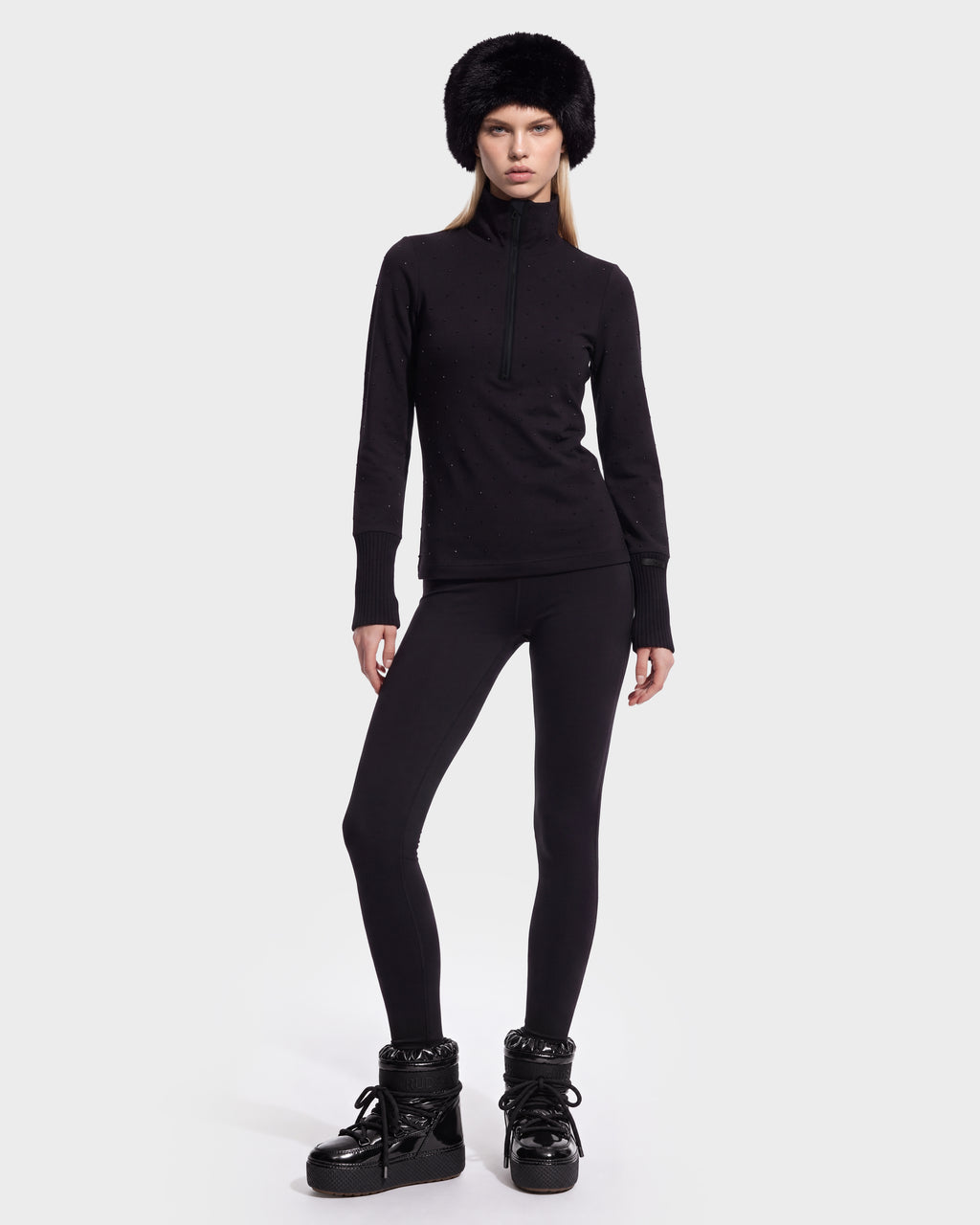 A person wears a black NEVA - 8526524 Black quarter-zip long-sleeve top styled with black leggings, a faux-fur hat, and black winter boots.