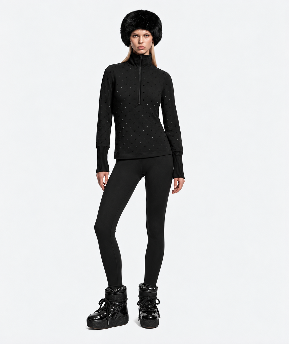 A person wearing a black faux-fur hat, a NEVA - 8526524 Black stretch nylon quarter-zip top with stud detailing, black leggings, and insulated boots for a refined cold-weather layering look.