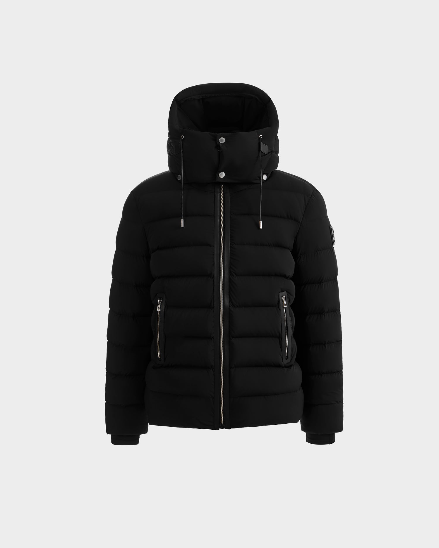 The Rudsak NAVAN - 6125598 Black is a black quilted puffer jacket with a high collar, hood, front zipper, and two zippered side pockets, shown against a plain light gray background.