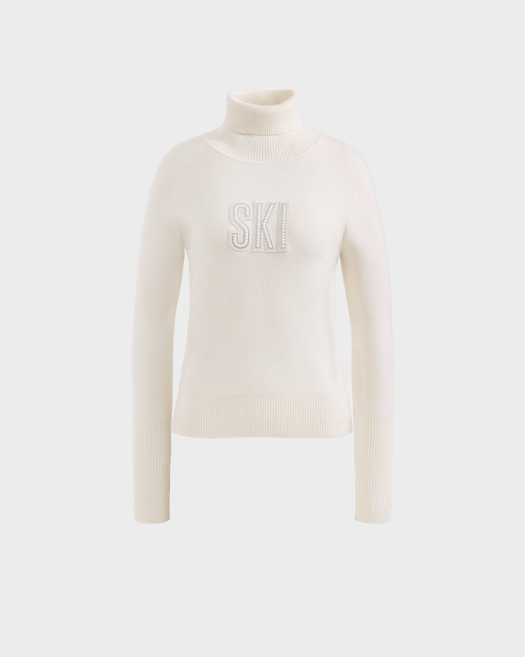The Rudsak NOVIE - 8525659 Vanilla is a women’s white knit turtleneck ski sweater with ribbed cuffs and hem, featuring bold, textured SKI lettering on the front, shown against a plain light background.