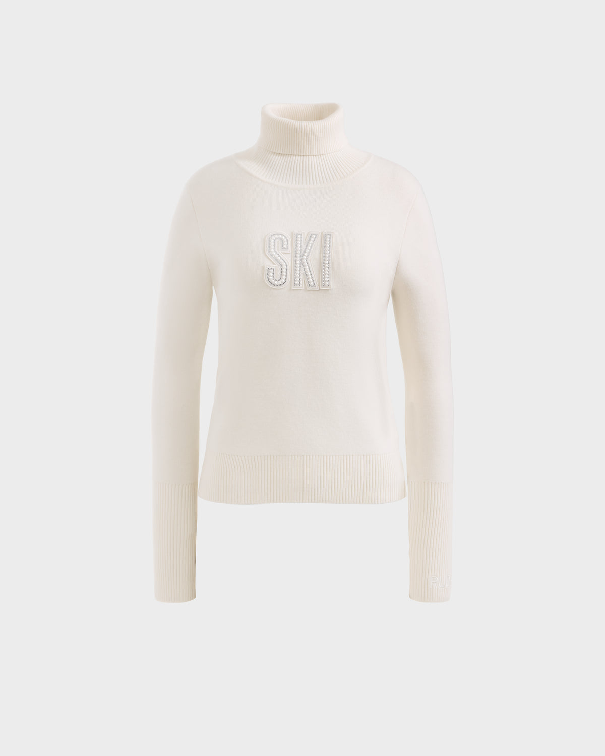 The Rudsak NOVIE - 8525659 Vanilla is a women’s white knit turtleneck ski sweater with ribbed cuffs and hem, featuring bold, textured SKI lettering on the front, shown against a plain light background.