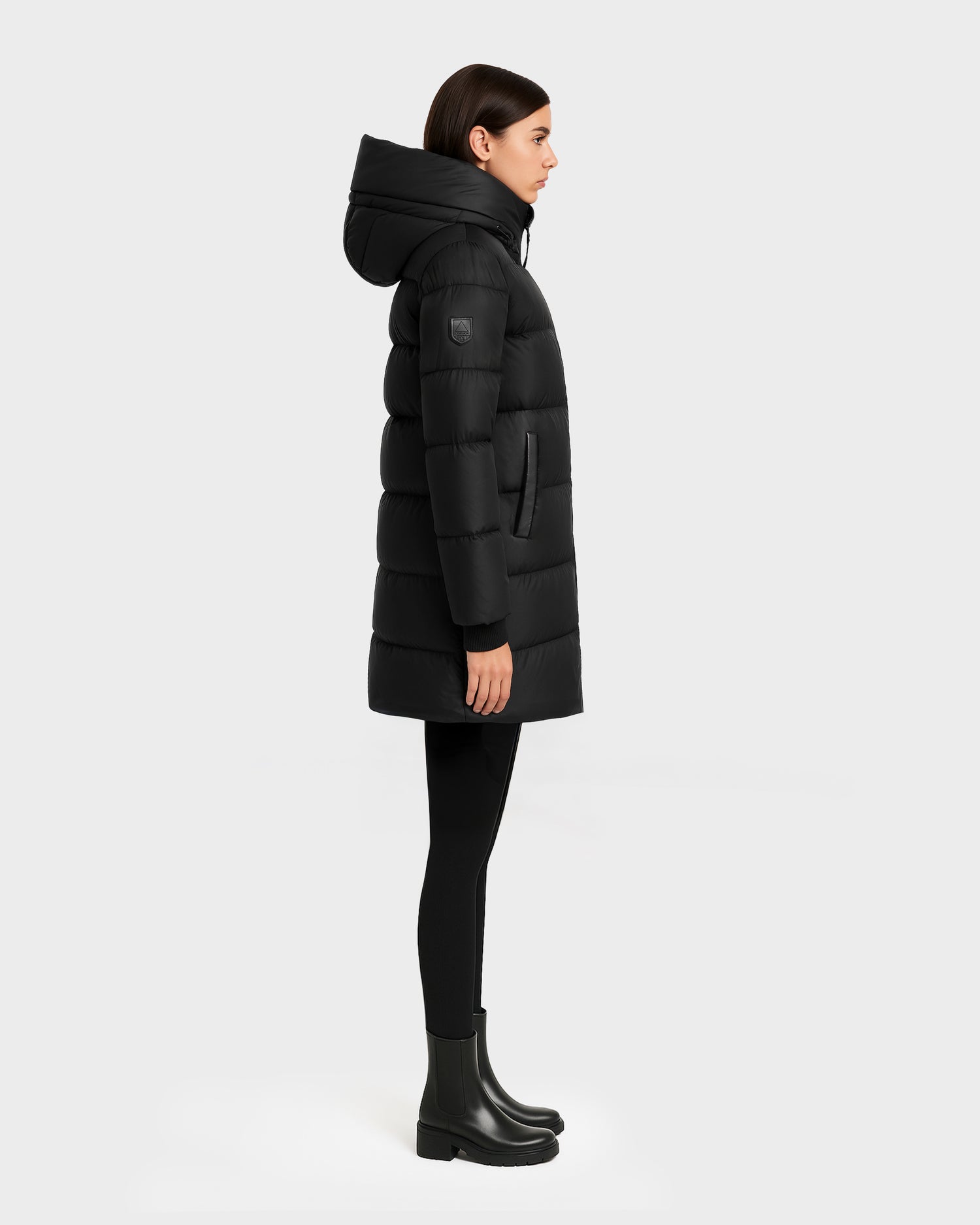 A woman stands in profile against a light gray background, wearing the Rudsak OLIVIA - 4123509 Black hooded puffer coat, paired with black leggings and black boots.