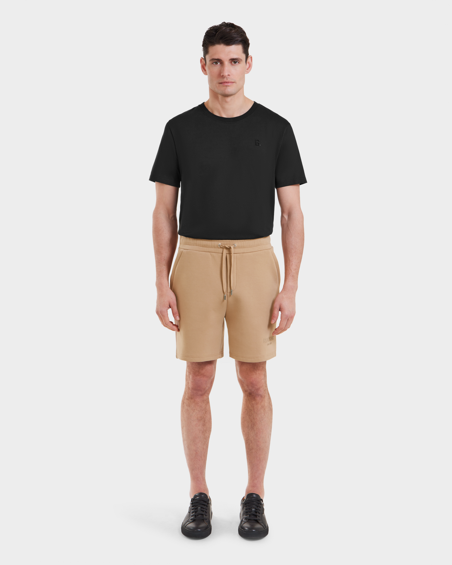 A man stands facing forward in a Rudsak PASCAL - 6524022 Almond t-shirt, casual drawstring shorts, and black sneakers against a plain white background.
