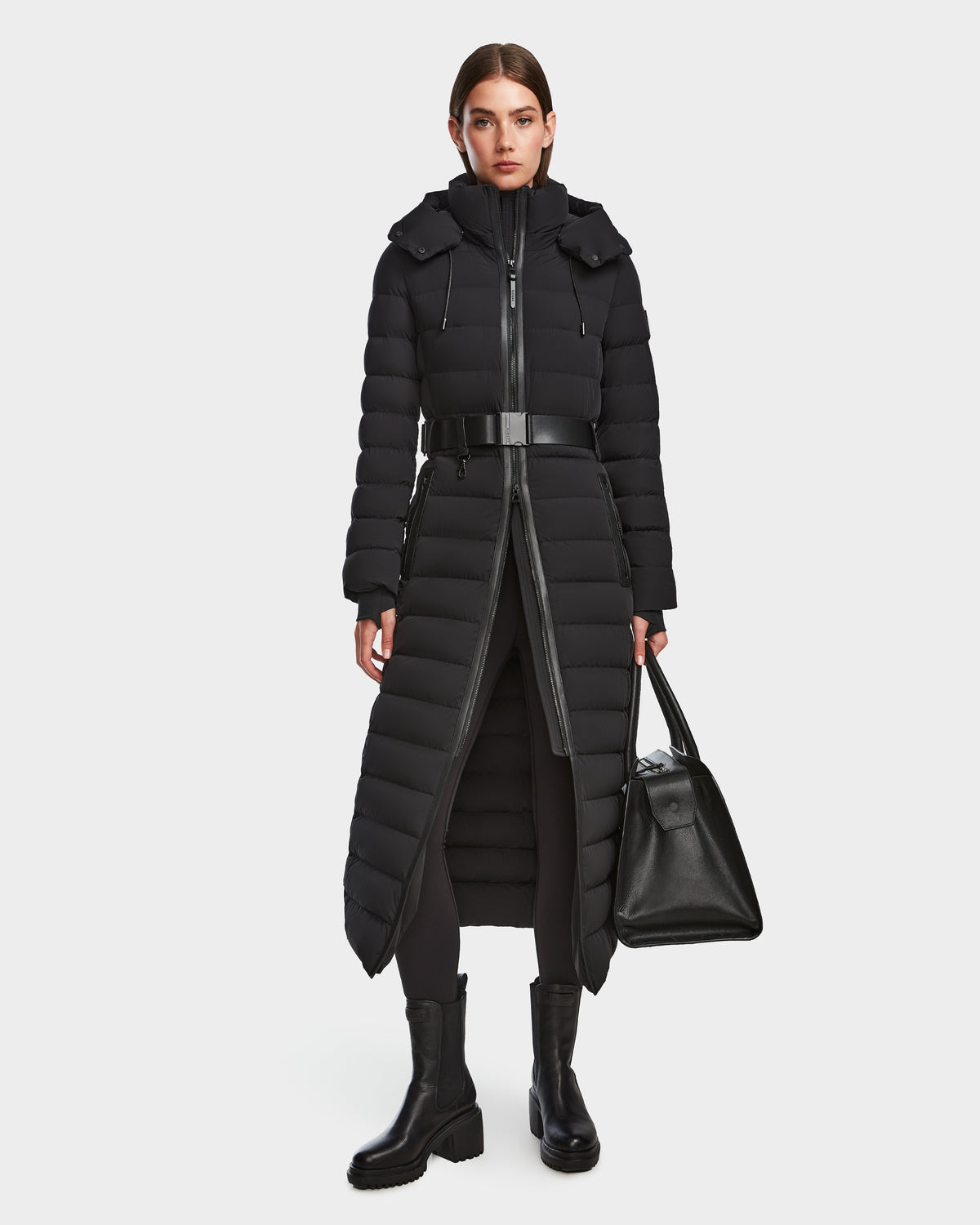 A woman stands facing forward against a plain white background wearing the Rudsak PELIN L - 8124815 Black womens long down puffer winter coat, paired with black pants, chunky black boots, and holding a large black handbag.