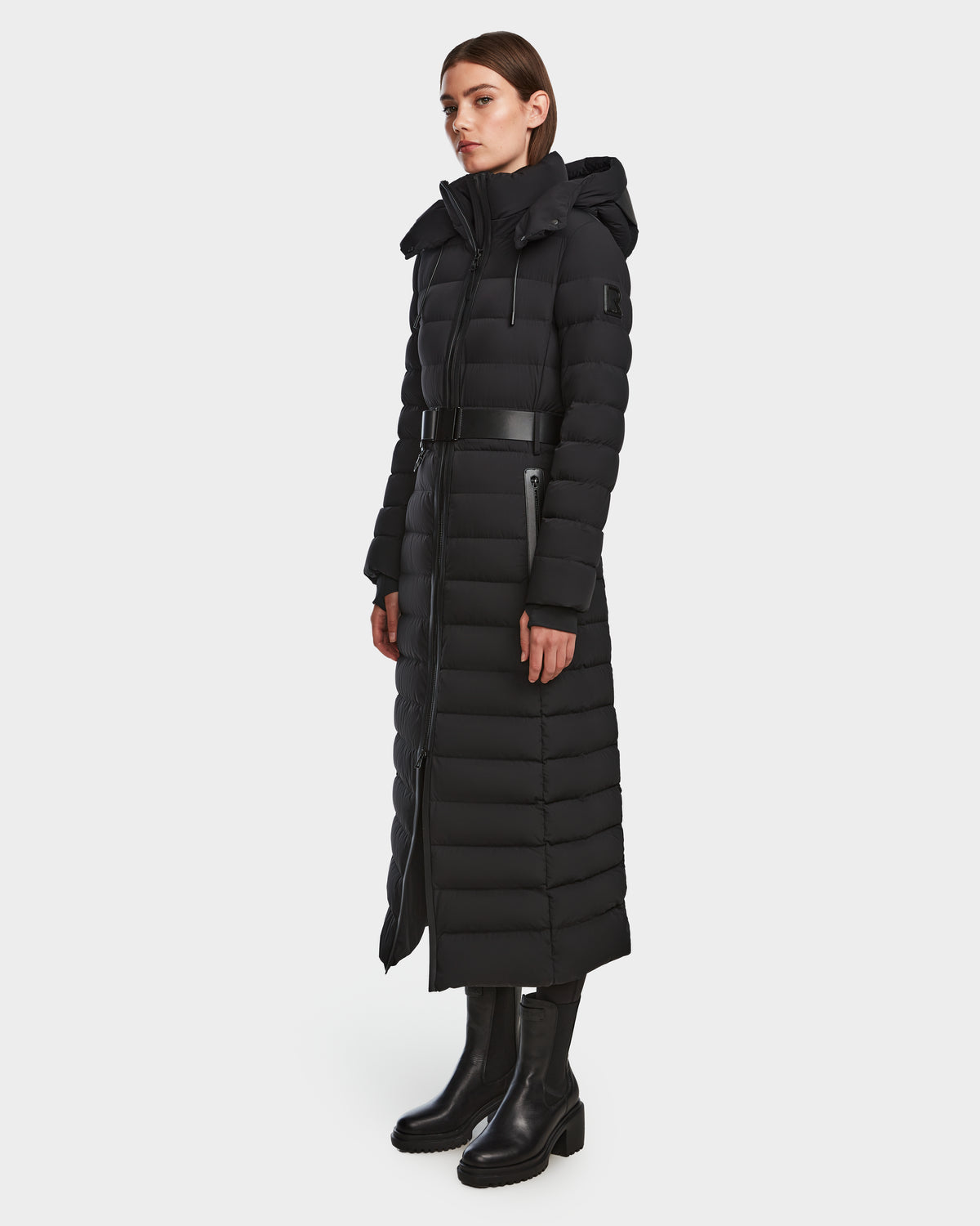 A woman wears the Rudsak PELIN L - 8124815 Black womens long down puffer winter coat with a belt, front zipper, hood, and black boots, crafted with RDS-certified down, set against a plain white background.