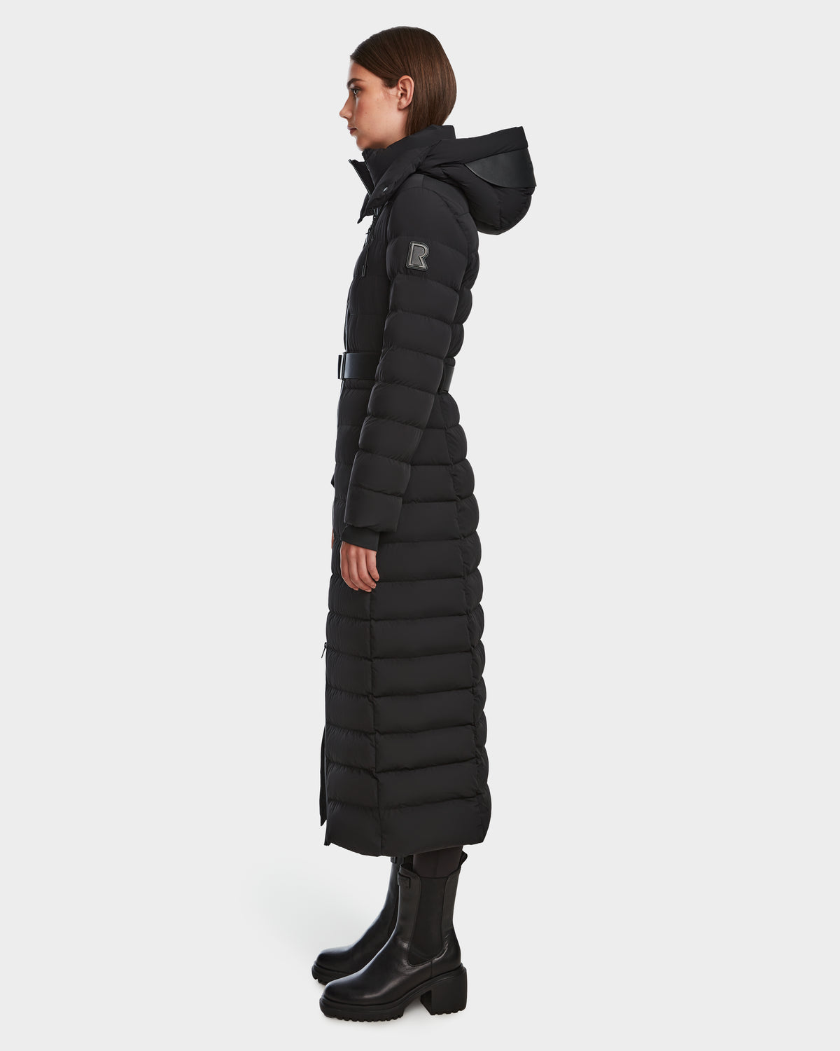 A woman stands in profile on a plain white background wearing the Rudsak PELIN L - 8124815 Black womens long down puffer with hood, belted waist, horizontal quilting, RDS-certified down, and black boots. The coat is mid-calf length.