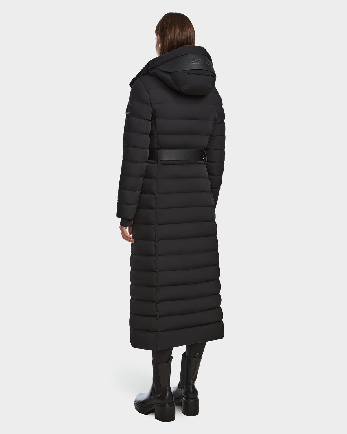 A person wearing the Rudsak PELIN L - 8124815 Black womens long down puffer coat with a belt and hood up stands with their back to the camera against a plain white background.