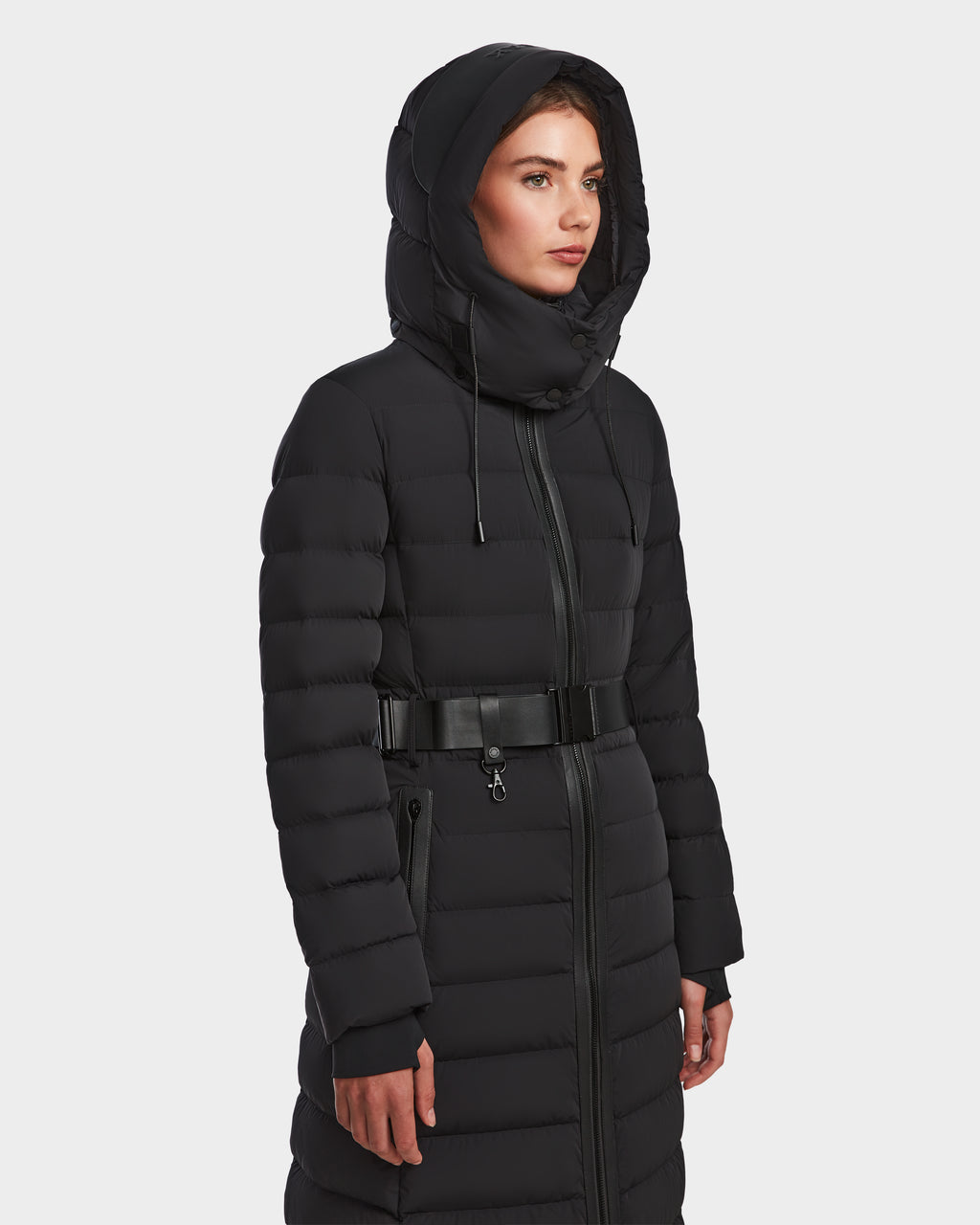 A woman models the Rudsak PELIN L - 8124815 Black womens long down puffer winter coat with hood and belted waist, standing slightly to the right against a plain white background.
