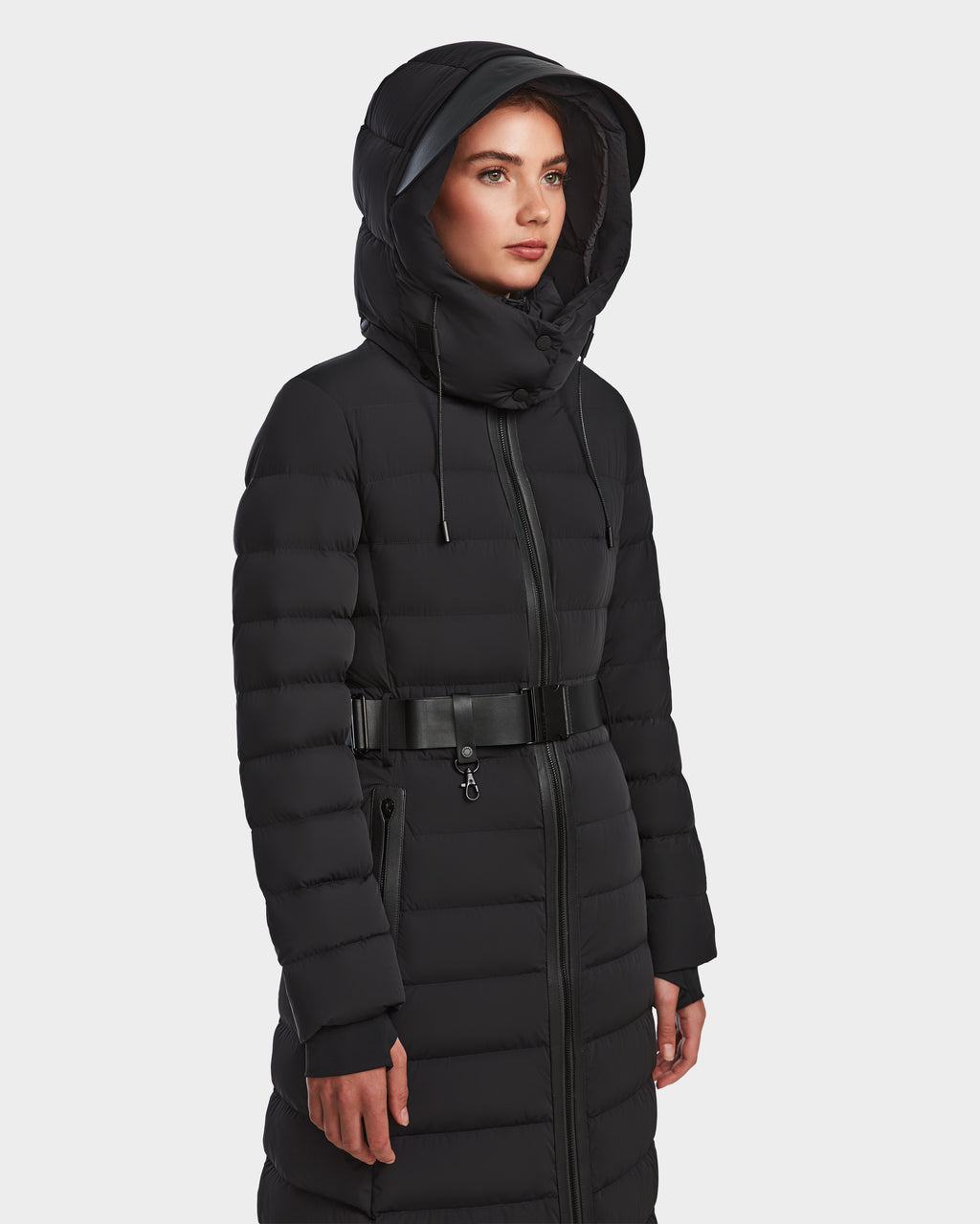 A woman wearing the Rudsak PELIN L - 8124815 Black long down puffer—with hood and belt—stands against a white background, facing slightly right. This stylish winter coat is crafted with RDS-certified down for warmth and comfort.
