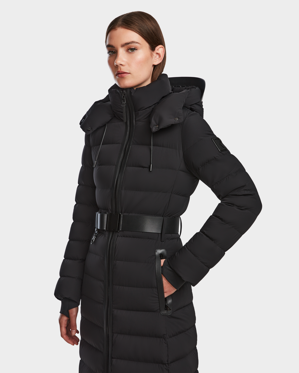 A woman wears the Rudsak PELIN L - 8124815 Black women’s long down puffer coat with a high collar and hood, standing confidently against a plain white background.