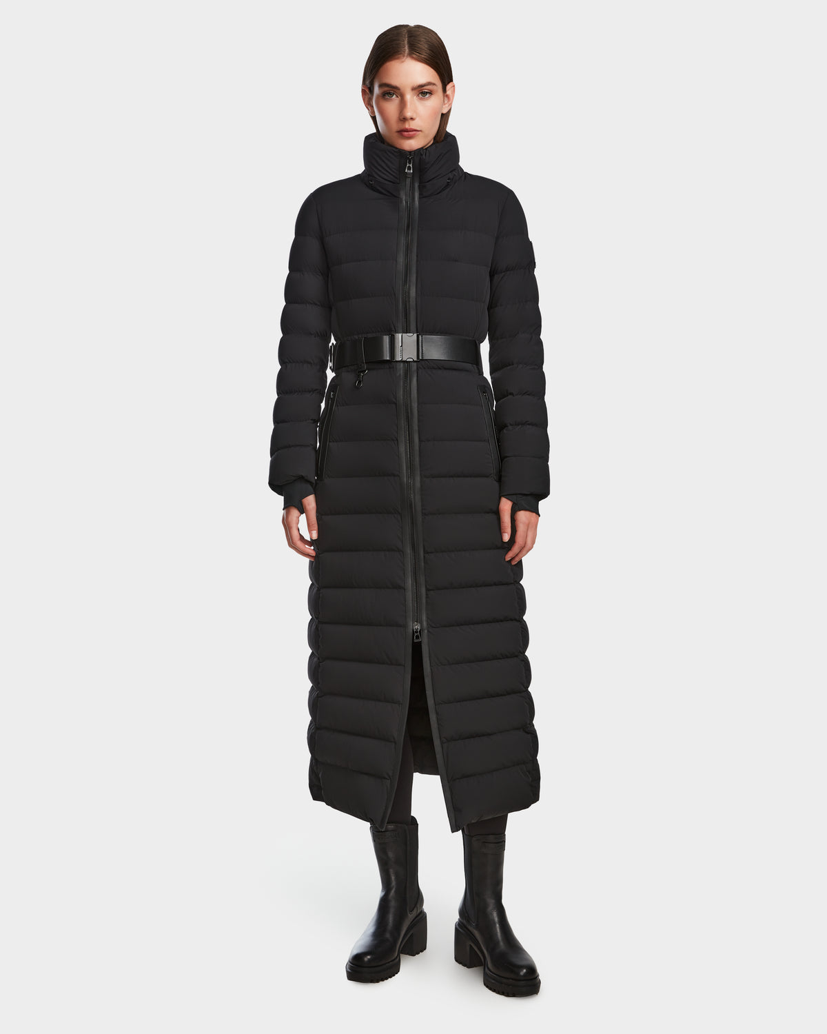 A woman faces forward in a Rudsak PELIN L - 8124815 Black womens long down puffer coat with high collar, belted waist, and front zipper, paired with black boots. The RDS-certified down keeps her warm. Background is plain white.