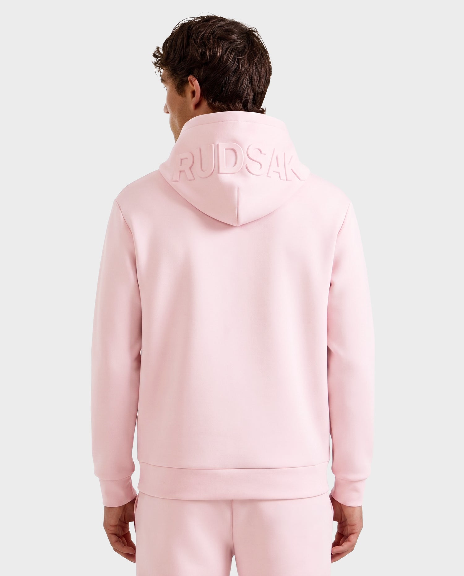 A person is seen from the back wearing the Rudsak LEROY - U525509 hoodie and matching pants in light pink, with "RUDSAK" embossed on the hood, set against a plain light-colored background.