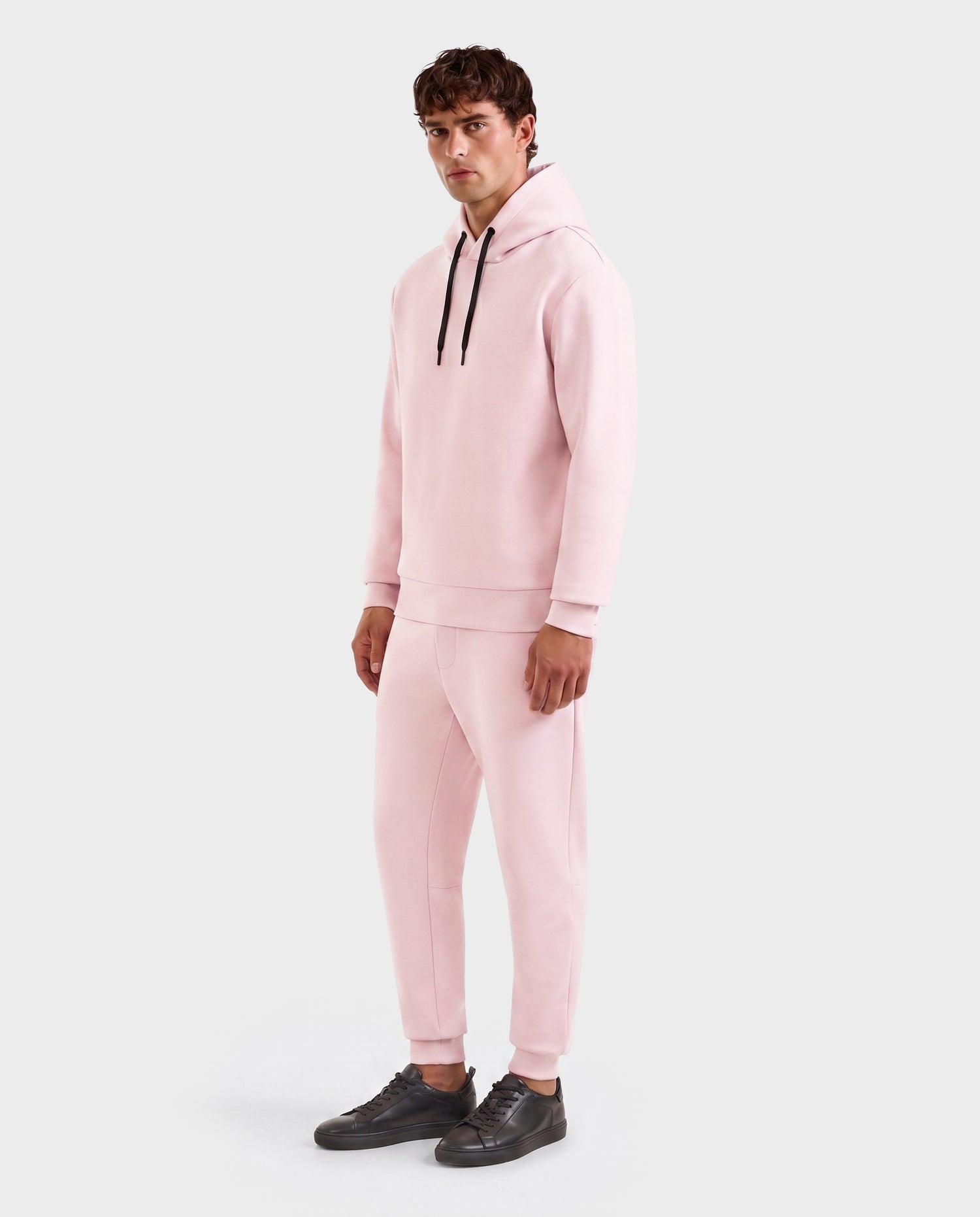 A person wears the Rudsak LEROY - U525509 Light Pink M gender-inclusive hoodie with black drawstrings and matching sweatpants, paired with black sneakers, against a plain white background.