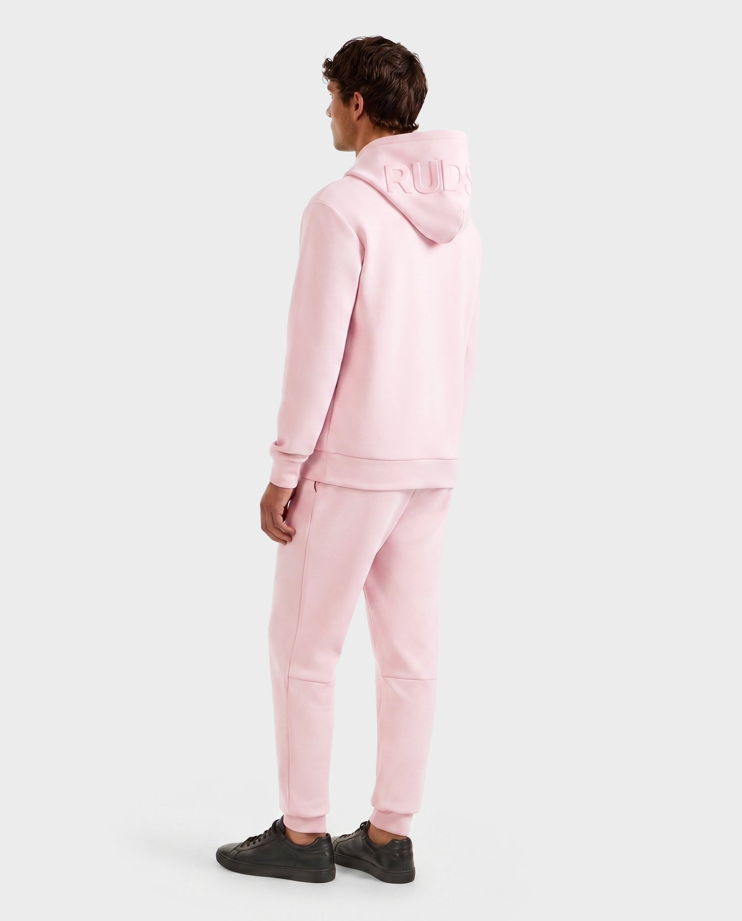A person faces away, wearing the LEROY - U525509 Light Pink M unisex hoodie and sweatpants by Rudsak with black sneakers. The hood features embossed text. The background is plain white.
