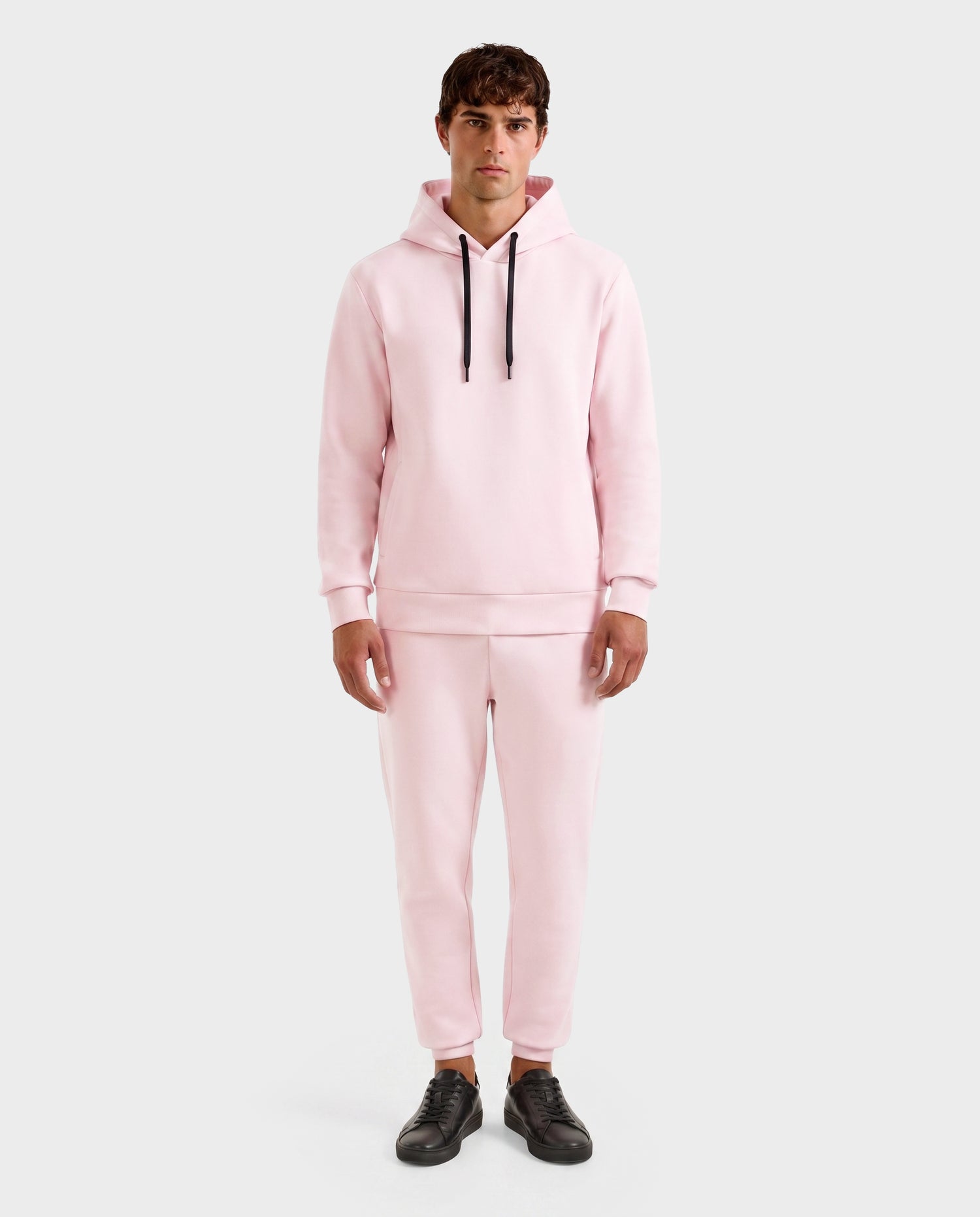 A person stands facing forward in the Rudsak LEROY - U525509 Light Pink M gender-inclusive hoodie and jogger pants with black drawstrings, black sneakers, against a plain light gray background.