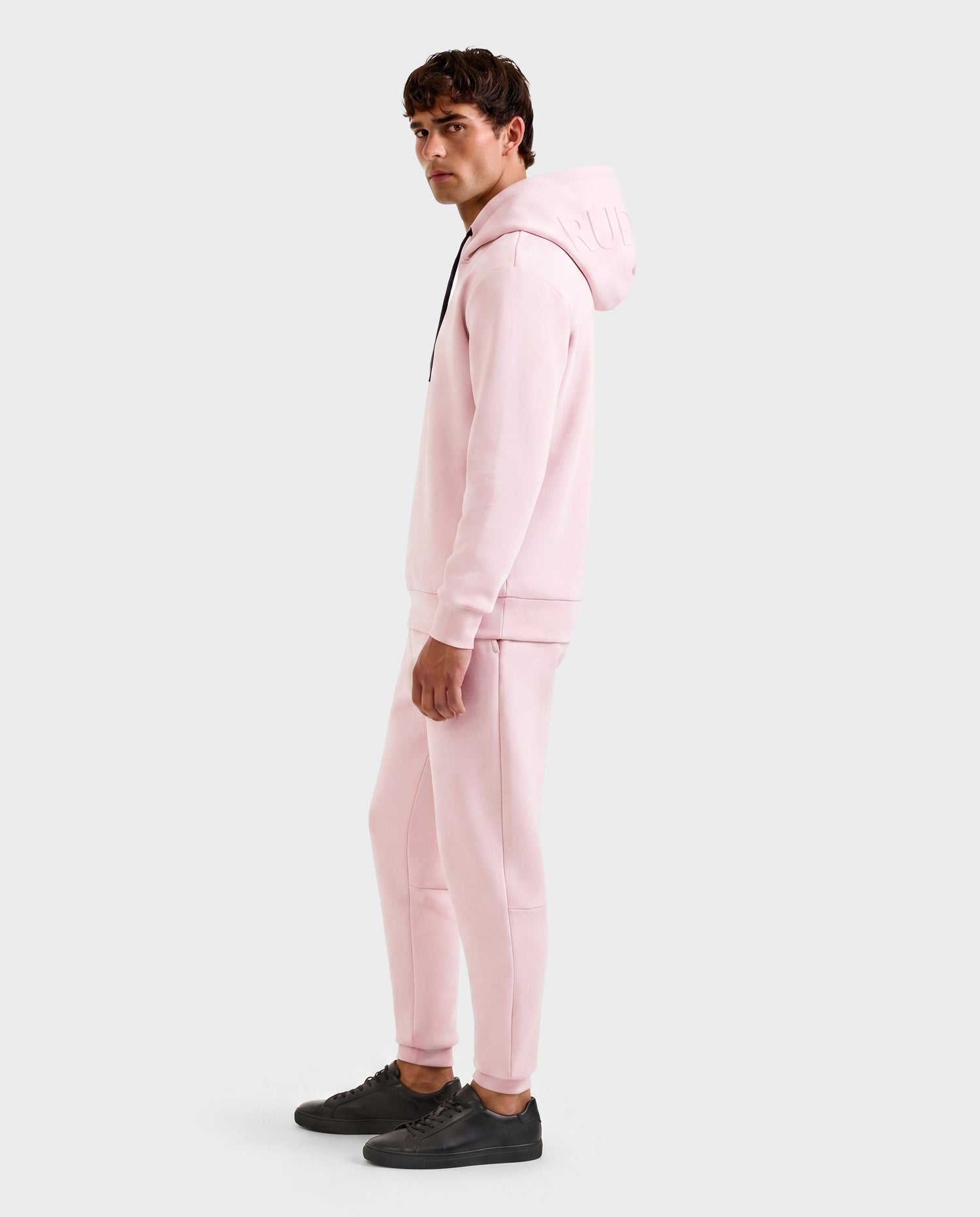 A man stands sideways, facing the camera, wearing the Rudsak LEROY - U525509 Light Pink M gender-inclusive hoodie with hood up and matching pink joggers. He pairs them with black sneakers against a plain white background.
