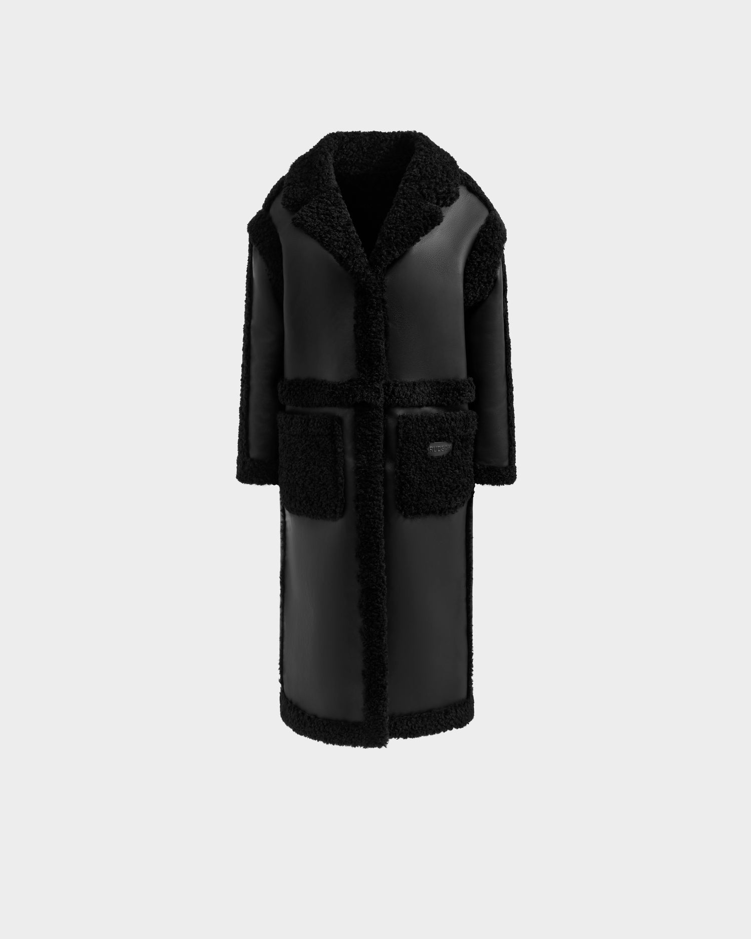 The Rudsak PALOMA - 8125675 Black is a long faux shearling coat with a textured fuzzy lining, wide collar, two large front pockets, and a relaxed straight fit, shown against a plain light gray backdrop.