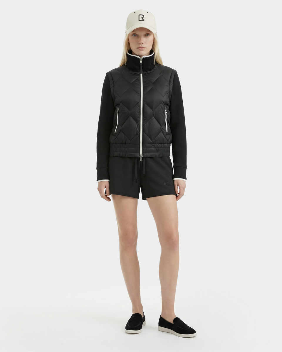 A woman faces forward against a white background, wearing the Rudsak PRISA - 8126022 Black convertible down jacket with removable sleeves, black shorts, slip-on shoes, and a beige cap with an "R" logo.