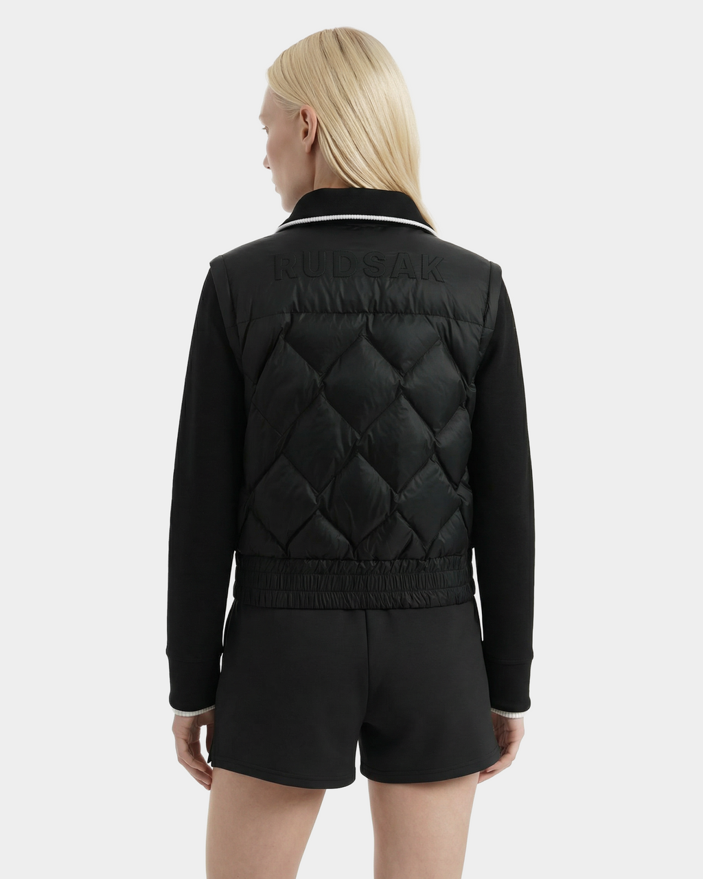 A person with straight blonde hair wears the Rudsak PRISA - 8126022 Black convertible down jacket, featuring RUDSAK embossed on the back, and black shorts, standing against a plain light gray background.