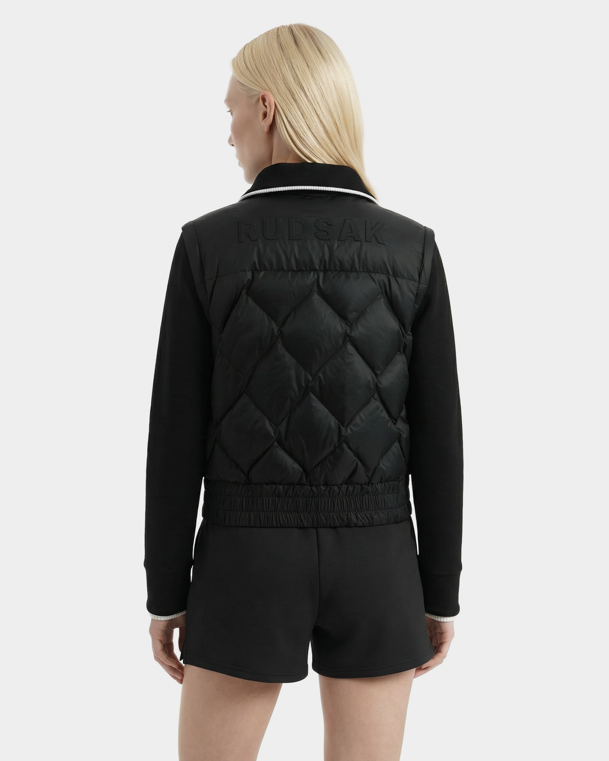 A person with straight blonde hair wears the Rudsak PRISA - 8126022 Black convertible down jacket, featuring RUDSAK embossed on the back, and black shorts, standing against a plain light gray background.