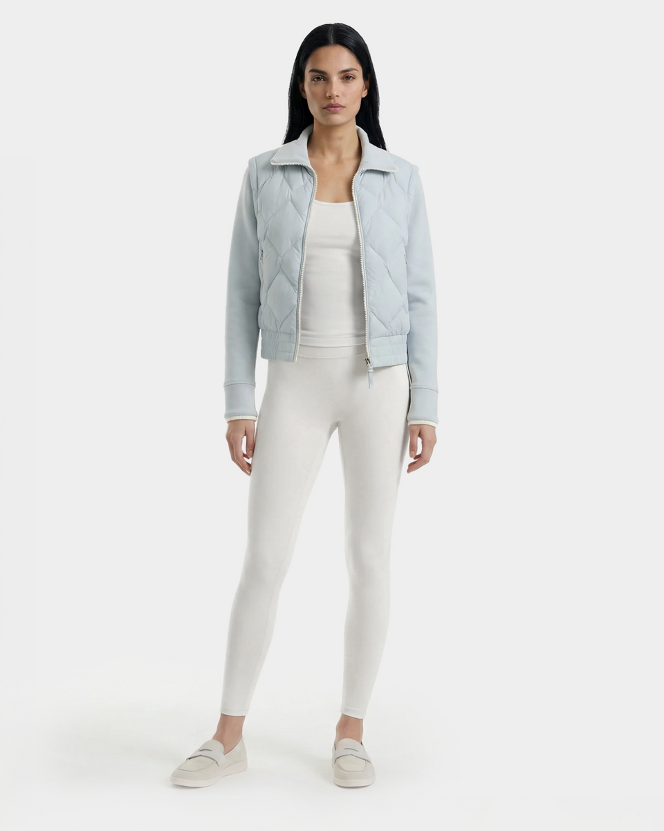 A woman faces forward wearing the Rudsak PRISA - 8126022 Powder Blue convertible down jacket with removable sleeves over a white top and leggings, paired with light slip-on shoes, against a plain white background.