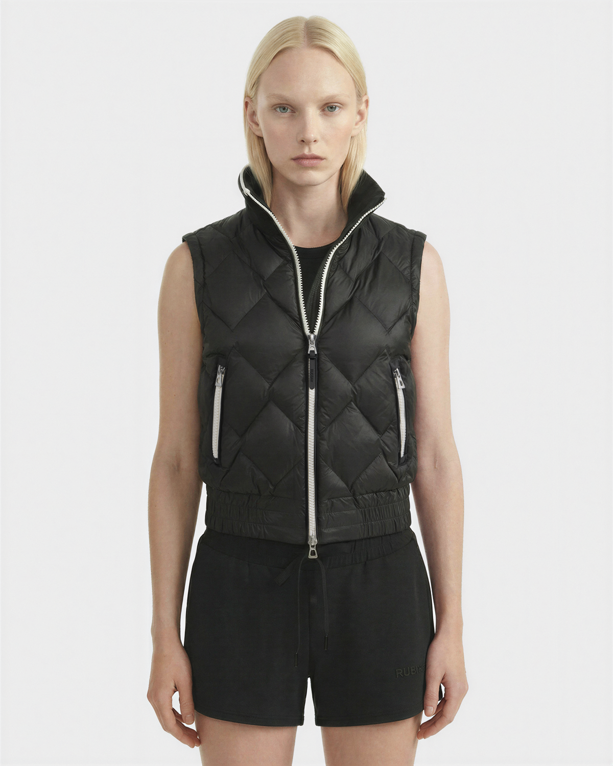 A person with straight blonde hair wears the Rudsak PRISA - 8126022 Black, a sleeveless quilted vest with zippered pockets and removable sleeves, paired with black shorts against a plain white background.