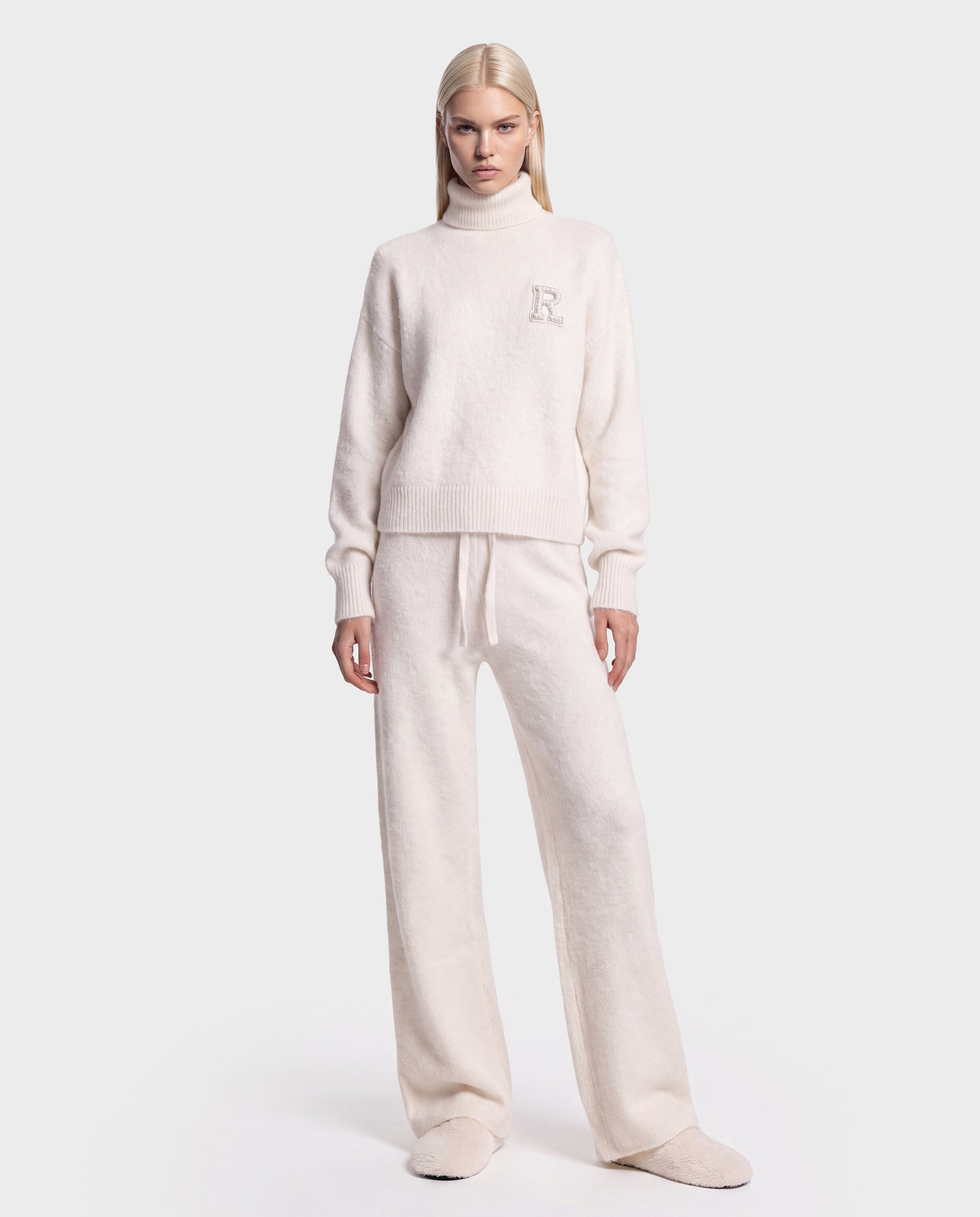 A person stands facing forward, wearing the Rudsak ALISYA - 8526505 Iceland cream-colored fuzzy knit turtleneck sweater and matching wide-leg pants with slippers, against a plain light background.