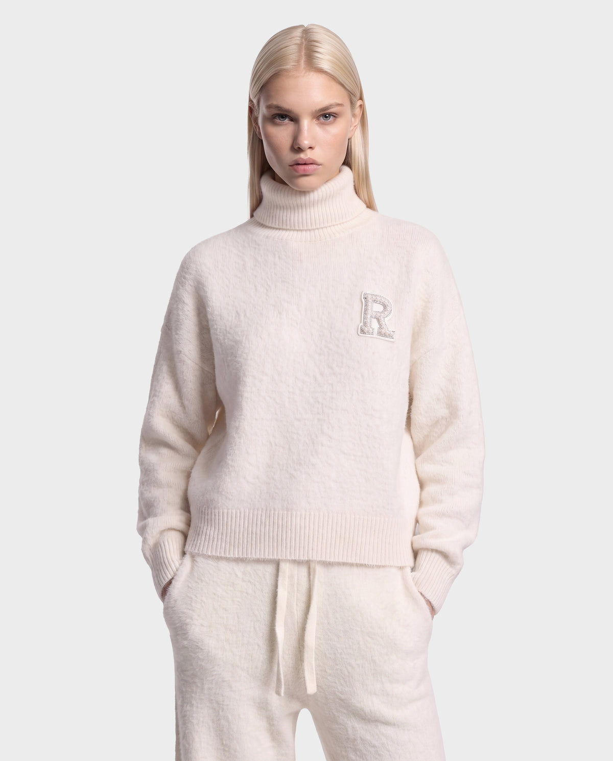 A person with long blonde hair wears the Rudsak ALISYA - 8526505 Iceland fuzzy knit cream turtleneck sweater featuring an R emblem, paired with matching cream pants, standing with their hands in their pockets against a plain light background.