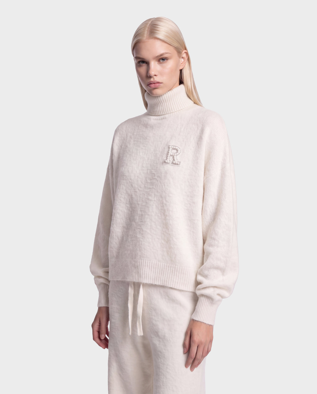 A person with long, straight blonde hair wears the cream-colored fuzzy knit ALISYA - 8526505 Iceland turtleneck sweater by Rudsak, featuring an “R” on the chest and matching drawstring pants with a relaxed silhouette, while standing against a plain light gray background.