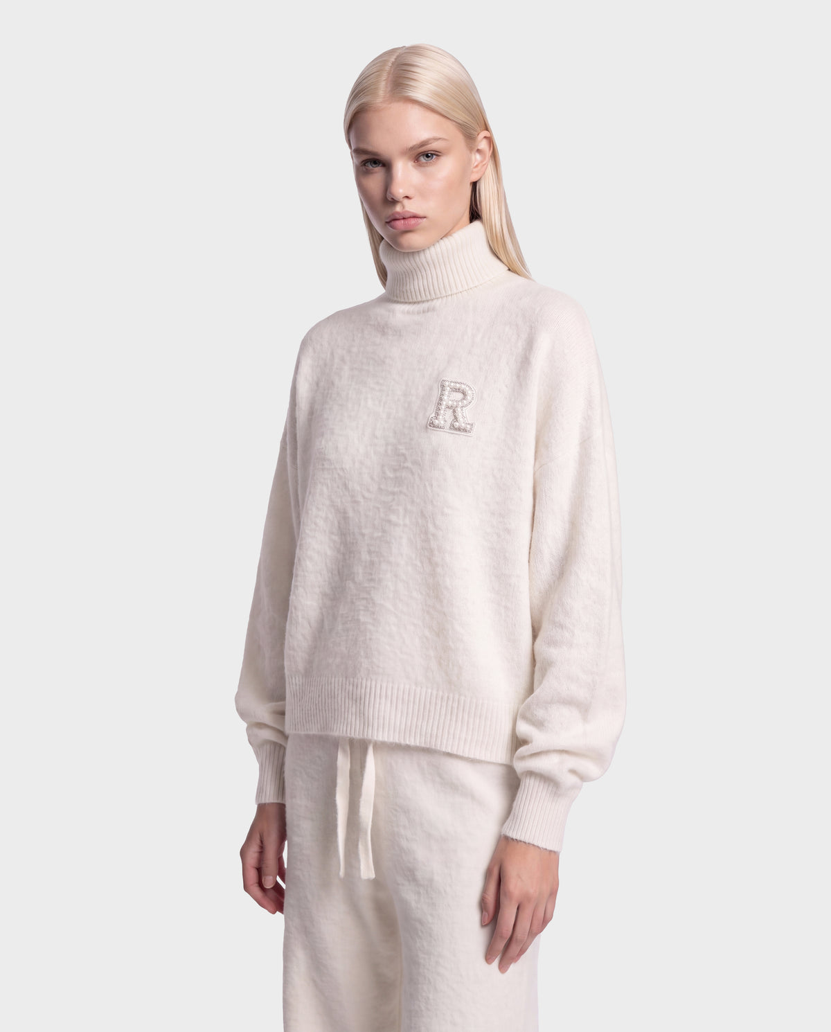 A person with long, straight blonde hair wears the cream-colored fuzzy knit ALISYA - 8526505 Iceland turtleneck sweater by Rudsak, featuring an “R” on the chest and matching drawstring pants with a relaxed silhouette, while standing against a plain light gray background.