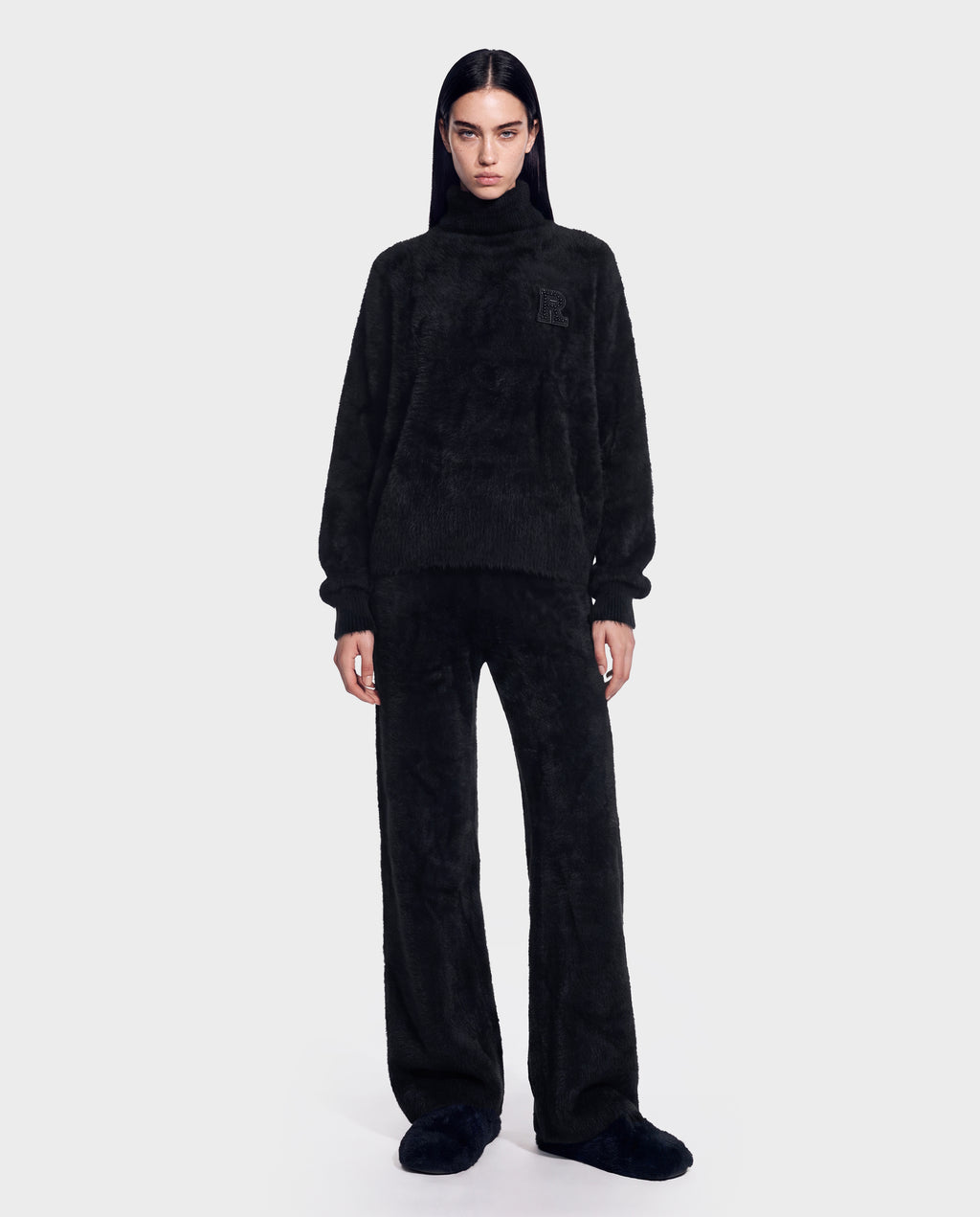 A person stands facing forward against a plain light gray background, wearing the Rudsak ALISYA - 8526505 Black hoodie and matching black pants in a relaxed, loose-fitting fuzzy knit silhouette, paired with black slippers.