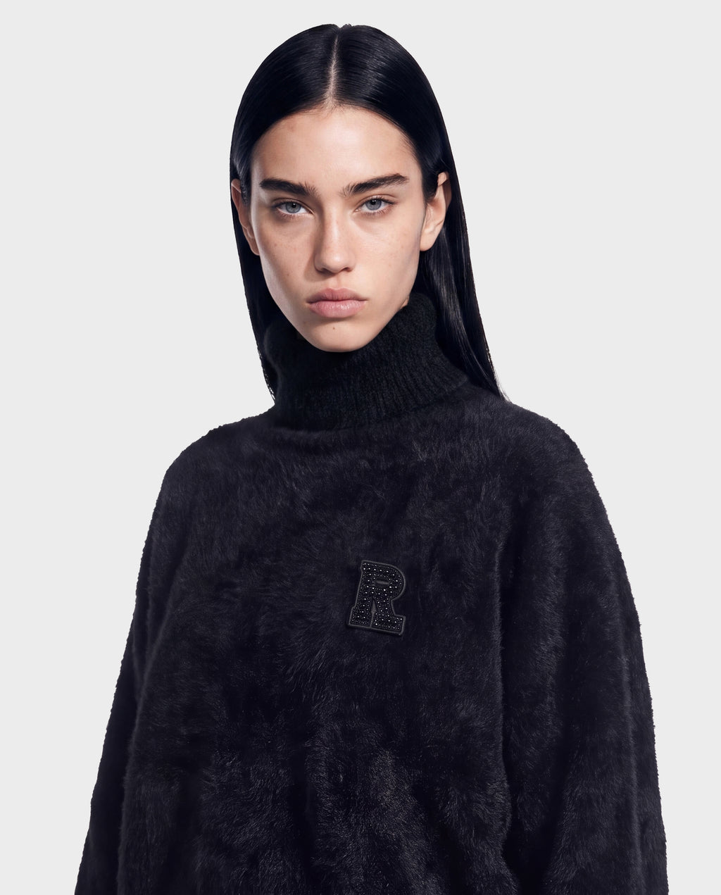 A person with long dark hair wears the ALISYA - 8526505 Black fuzzy knit turtleneck sweater by Rudsak, featuring a decorative letter R patch on the chest and a relaxed silhouette that stands out against a plain light background.