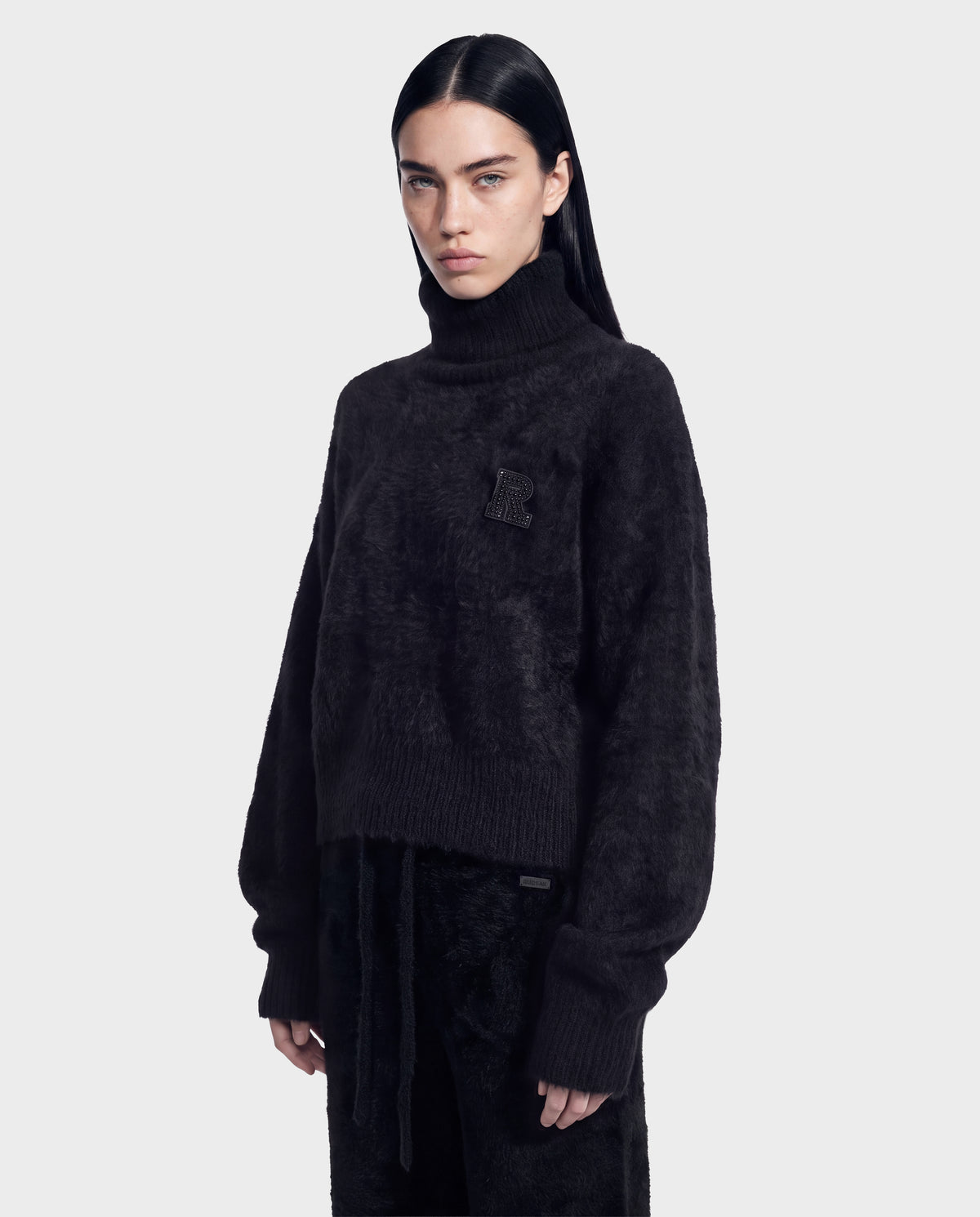 A person with long dark hair wears the Rudsak ALISYA - 8526505 Black fuzzy knit turtleneck sweater featuring a raised “R” patch on the chest, paired with black pants and standing against a light gray background.